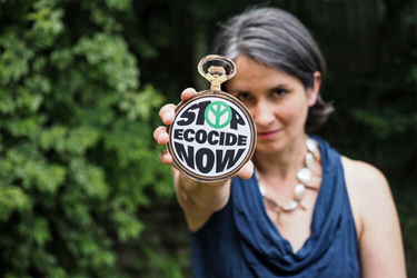 A picture of a woman holding a stopwatch to camera which says 'Stop Ecocide Now'
